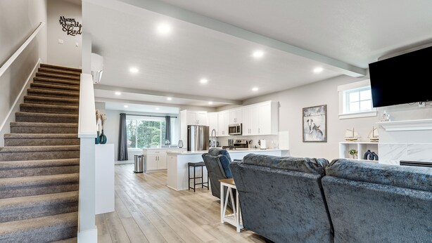 Una sala moderna con un sofá gris, una escalera y una zona de cocina con armarios blancos y un televisor de pantalla plana.