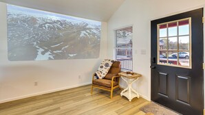 Een kamer met een projectiescherm waarop een bergachtig landschap te zien is, een houten stoel, een klein tafeltje met een schaal, en een donkere deur met een raam.