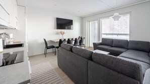 Un salon moderne avec un canapé d’angle gris, un téléviseur à écran plat, un coin repas avec des chaises et une grande fenêtre équipée de stores.
