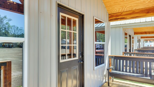A wooden porch with a bench, a door with a window, and a view of a parking lot. - 3 Mi to Sabine River: East Texas Cabin Hideaway (Joaquin)