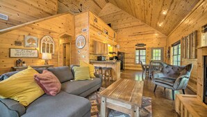 Een gezellig interieur van een blokhut met een grijze bank, een houten salontafel, en een eethoek met een bankje en stoelen.