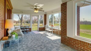 Una sala de estar con ventanales, ventilador de techo, mesa con sillas, sofá y vista a un área verde.