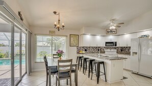 A modern kitchen with a large island, bar stools, a dining table with chairs, a ceiling fan, and a view of a swimming pool. - Cape Coral Family Home w/ Heated Pool (Cape Coral)