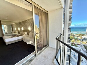 Une chambre d’hôtel comprenant un lit, un canapé, une petite table et une porte-fenêtre coulissante donnant sur un balcon avec vue sur l’océan et les bâtiments.