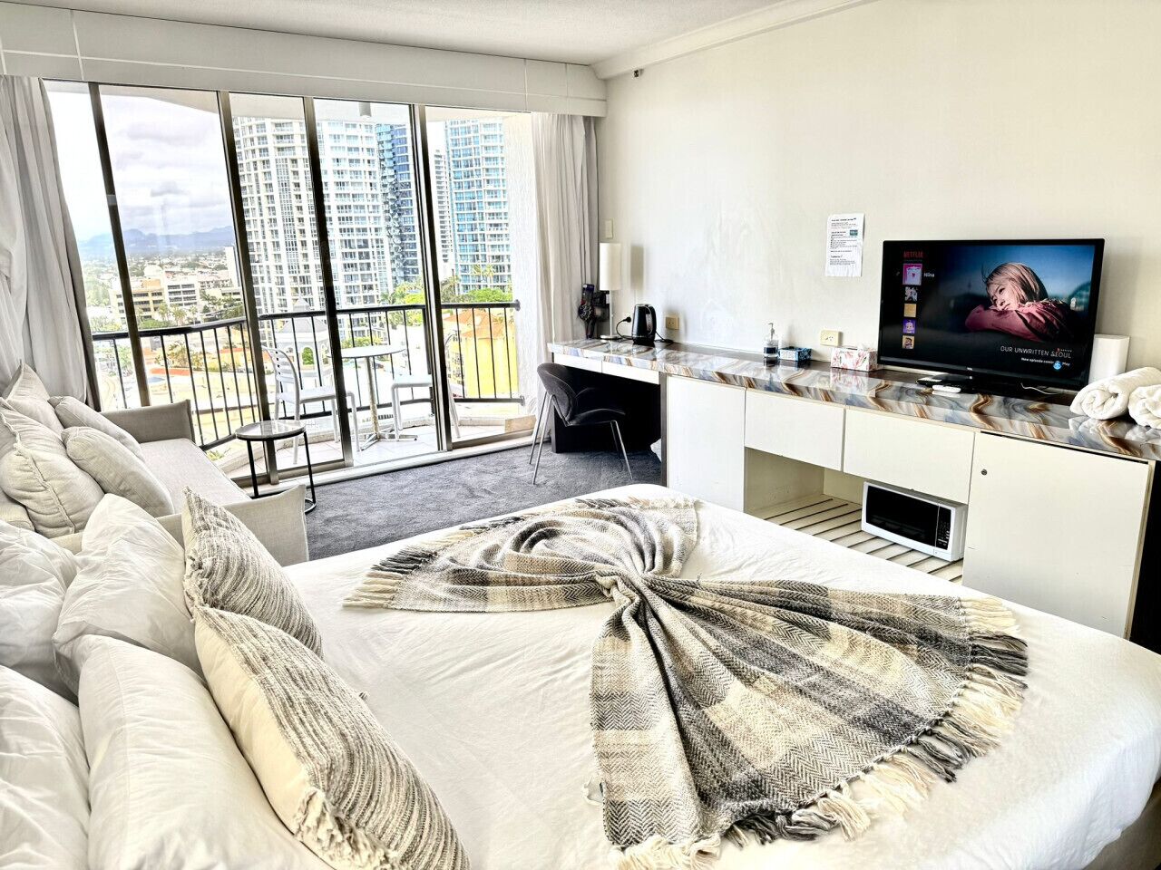 Ett hotellrum med en stor säng, ett skrivbord med en dator, en tv och en balkong med utsikt över staden.