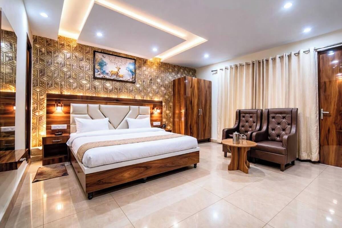 A modern bedroom with a large bed, a wooden wardrobe, a seating area with two chairs, and a decorative wall.
