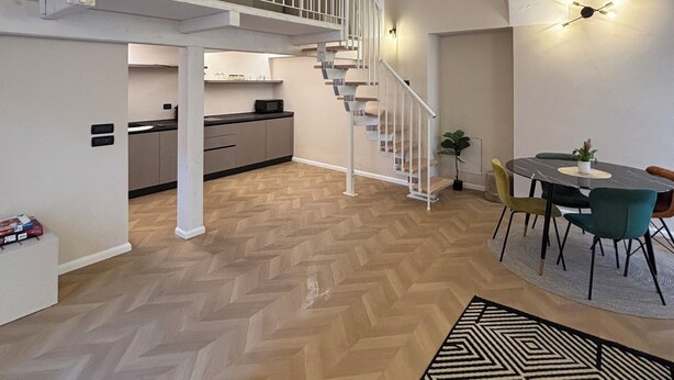 Un loft spacieux avec un parquet en point de Hongrie, un escalier doté d’une rampe blanche, un coin repas avec une table ronde et une cuisine à l’arrière-plan.