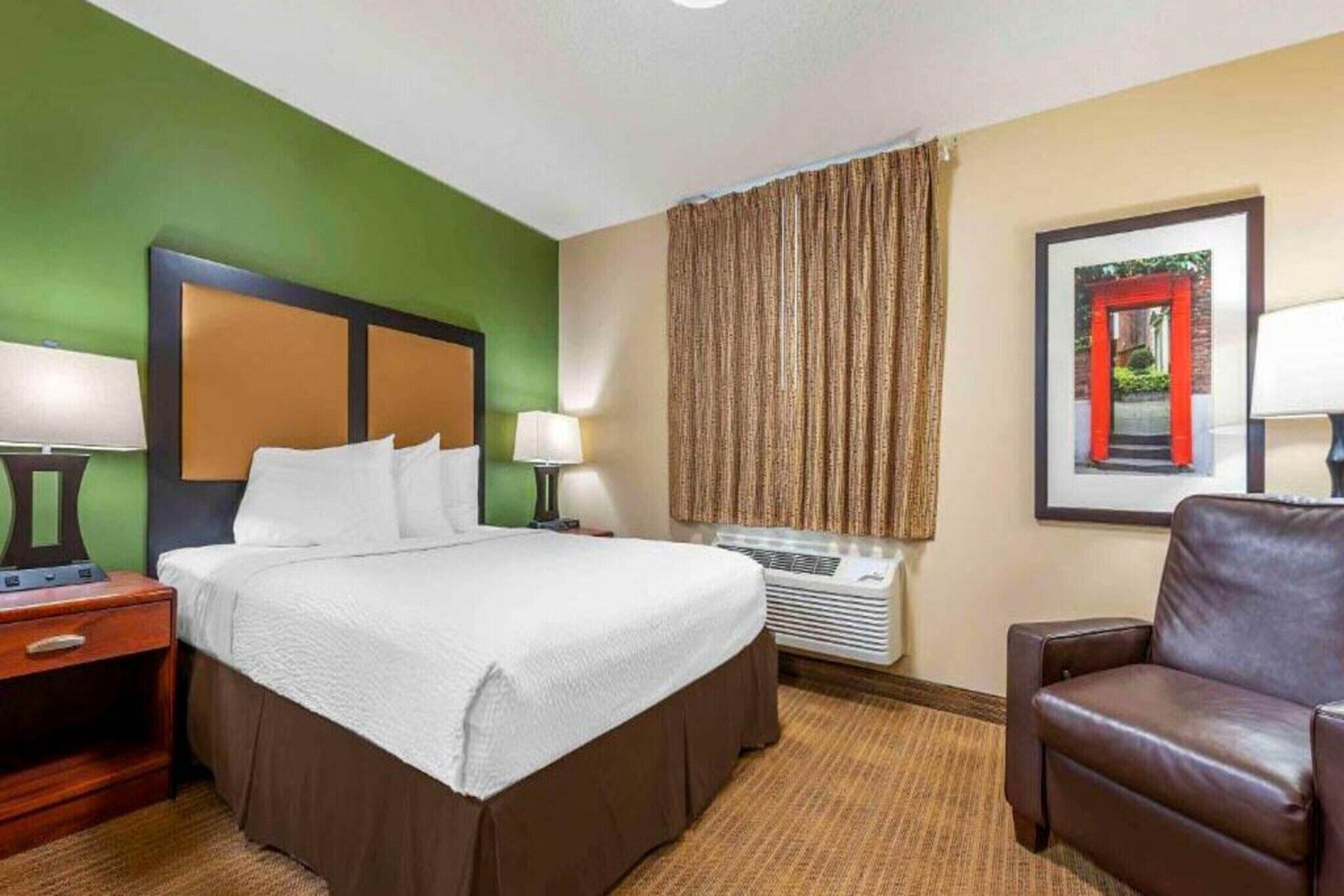 A hotel room with a bed, a desk, a chair, a window with curtains, and a framed picture on the wall.
