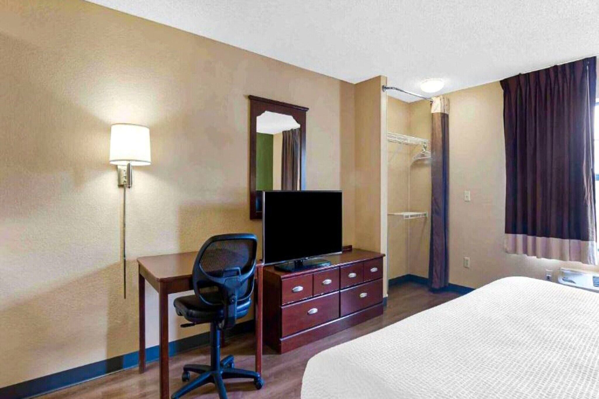 A hotel room with a bed, a desk with a chair, a television, a dresser, a mirror, and a shower.