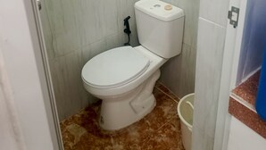 Un baño pequeño con inodoro blanco, barra para cortina de ducha y un balde en el suelo.