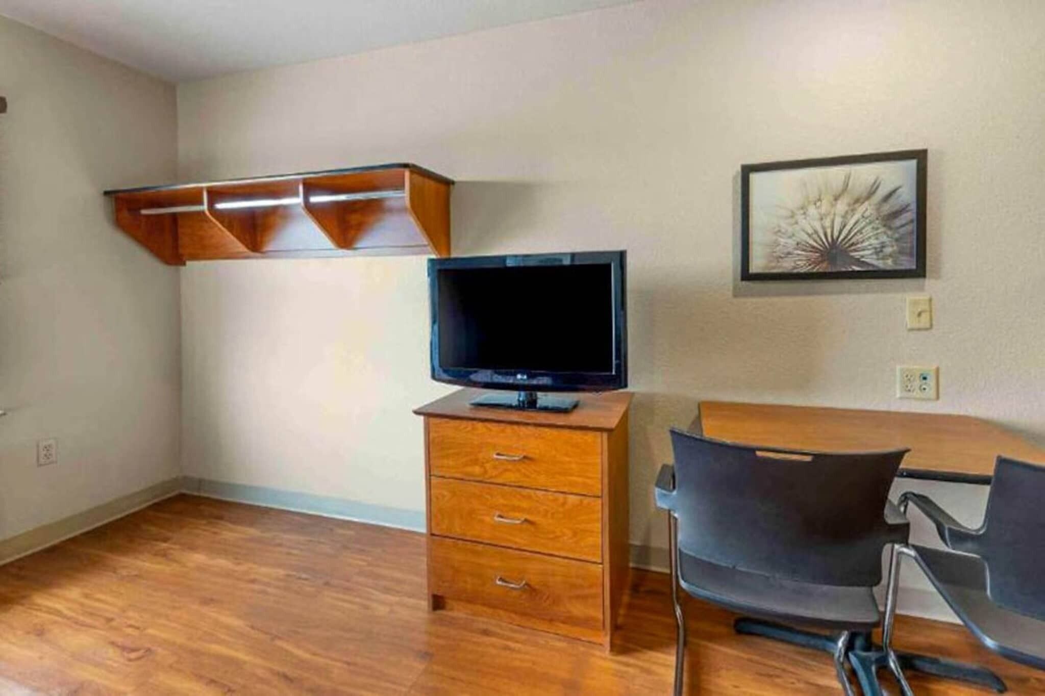 A room with a wooden dresser, a television on top, a dining table with chairs, and a framed picture on the wall.