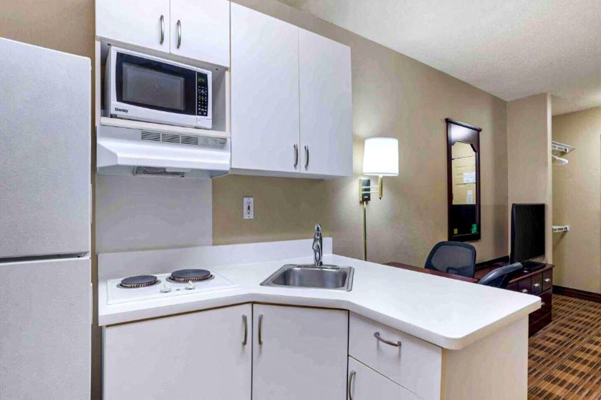 A compact hotel kitchen with a microwave, stove, sink, and a small seating area with a TV.