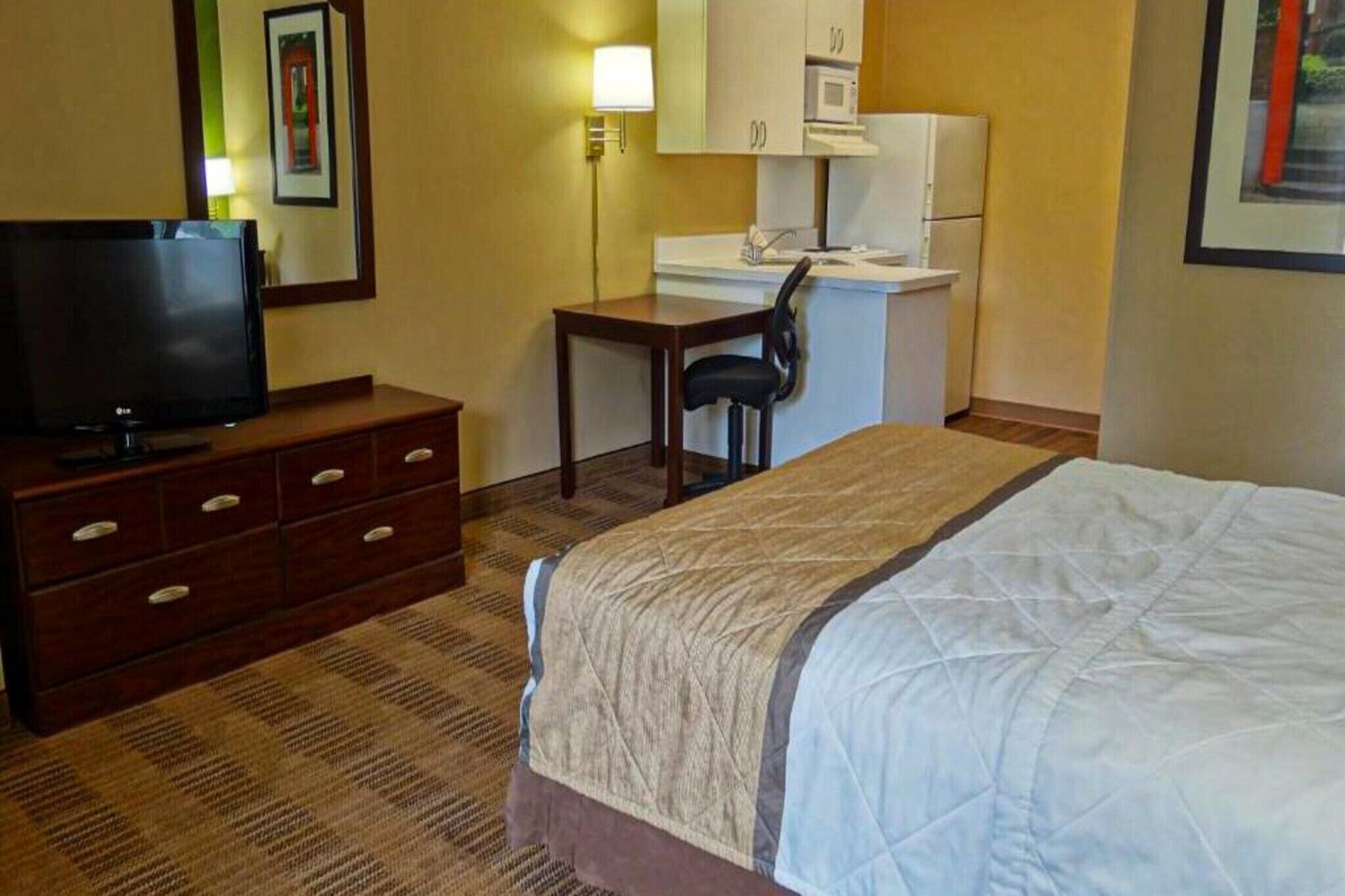 A hotel room with a bed, a TV, a desk with a chair, a microwave, and a small kitchenette.