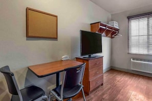A room with a wooden desk, two chairs, a television, a wall-mounted shelf with baskets, and a window with blinds. - 1 Bed | Deluxe, Sofa-Bed (Bossier City)