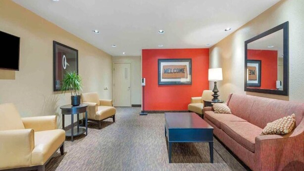 A hotel lobby with beige sofas, a coffee table, a red wall with a welcome sign, a potted plant, and a flat-screen TV.
