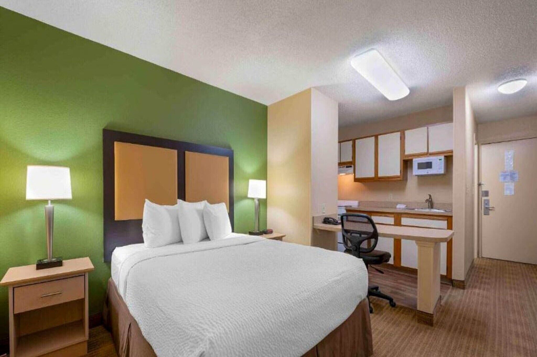 Ein Hotelzimmer mit Bett, Nachttischlampen, Schreibtisch, Stuhl und einer Küchenzeile.
