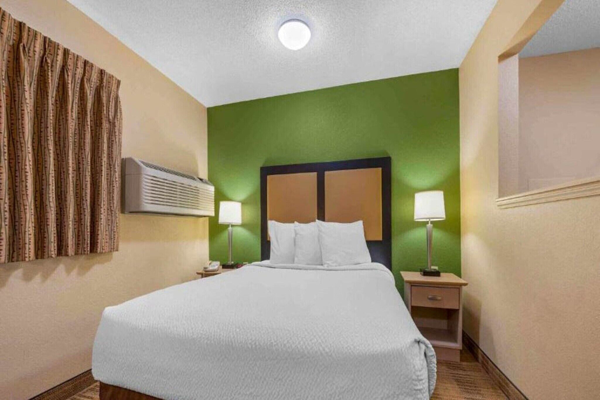 Habitación de hotel con cama, mesitas de noche, lámparas, equipo de aire acondicionado y una pared de acento verde.