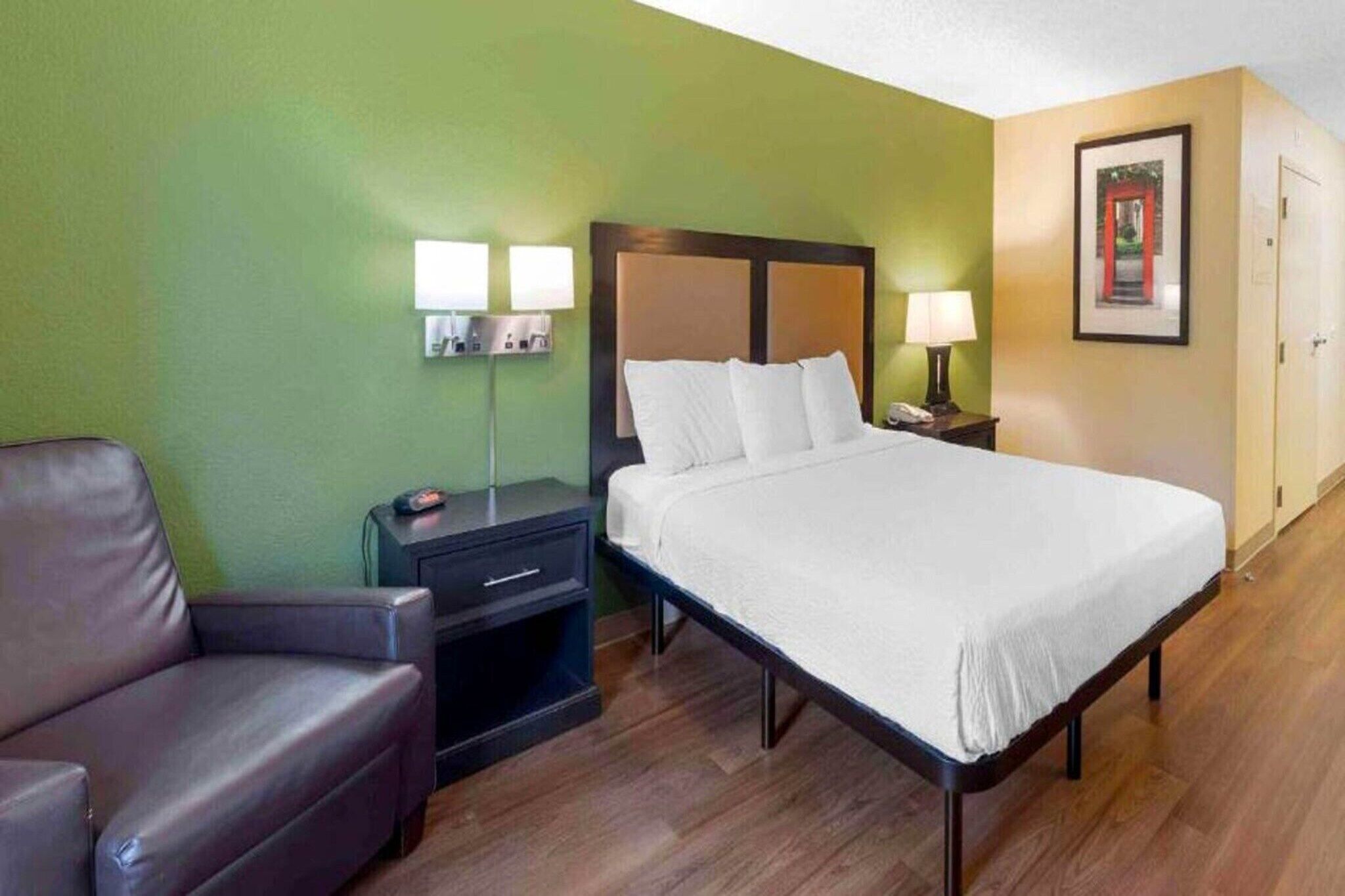 A hotel room with a bed, a nightstand, a lamp, a chair, and a framed picture on the wall.