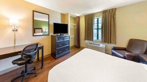 Ein Hotelzimmer mit Bett, Schreibtisch mit Stuhl, Fernseher, Kommode, Fenster mit Vorhängen und Heizung.