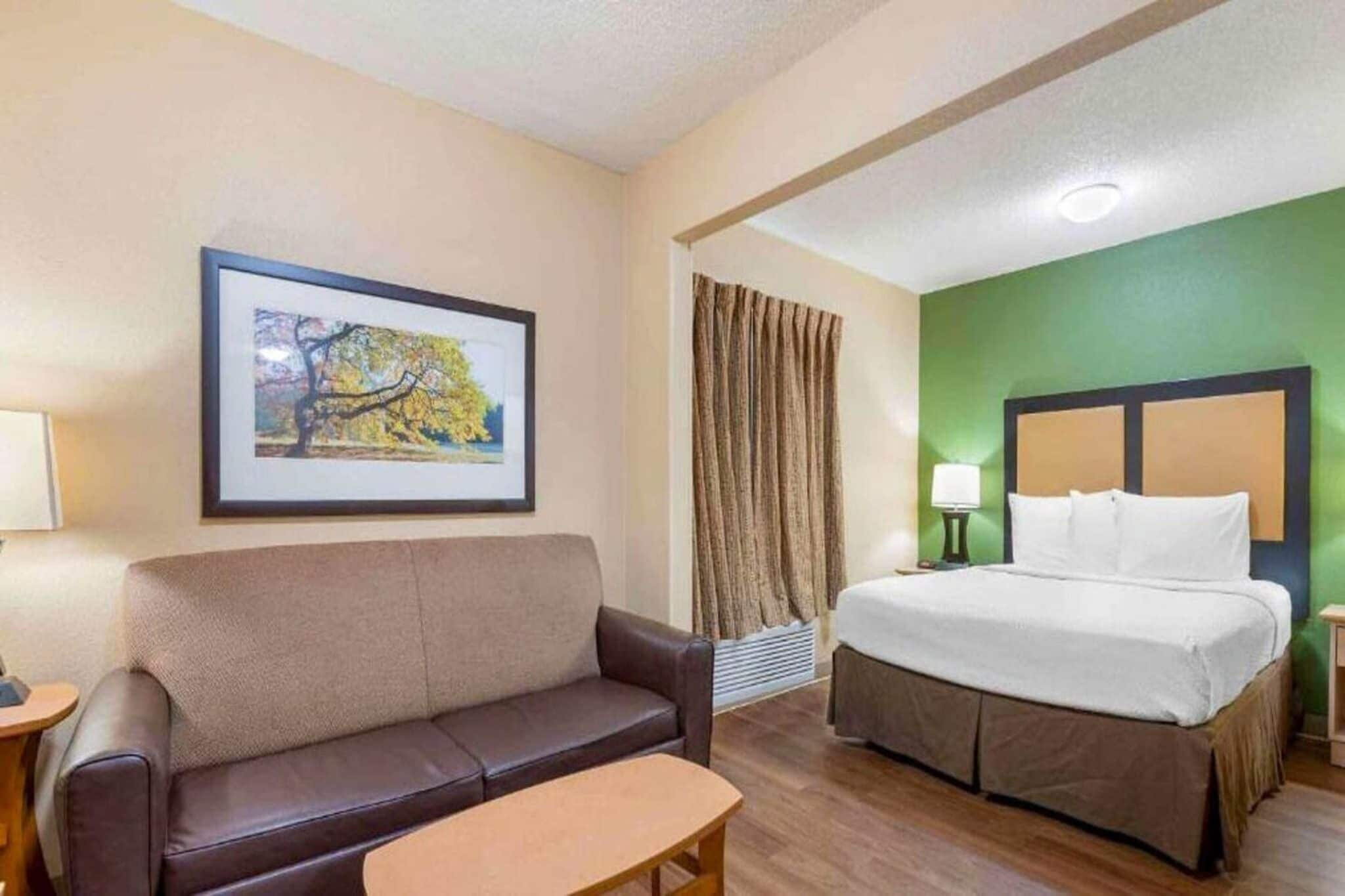 A hotel room with a bed, a couch, a small table, a framed picture on the wall, and a window with curtains.