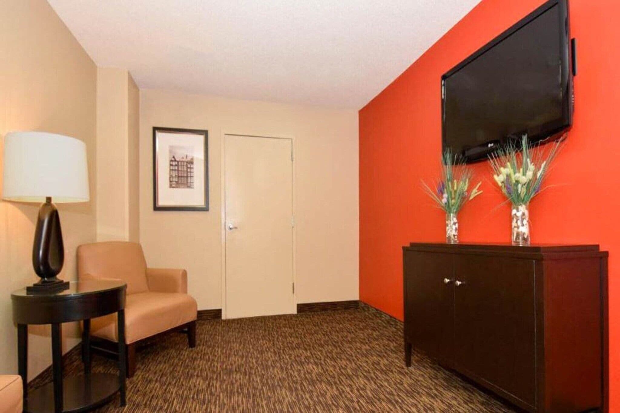 Ett hotellrum med en beige fåtölj, ett litet bord med en lampa, ett mörkt träskåp, en inramad tavla på väggen och en tv som är monterad på den orange väggen.