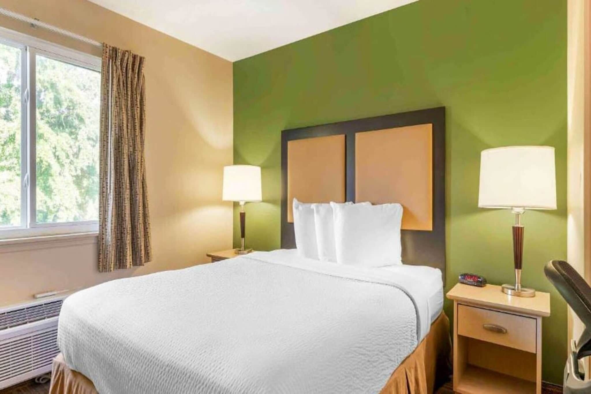 Ein Hotelzimmer mit einem Bett, zwei Nachttischlampen, einem Nachttisch, einem Fenster mit Vorhängen und einem an der Wand befestigten Kopfteil.