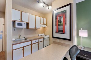 A compact kitchen with white cabinets, a microwave, a stove, and a sink. There's a framed picture on the wall and a desk with a chair.