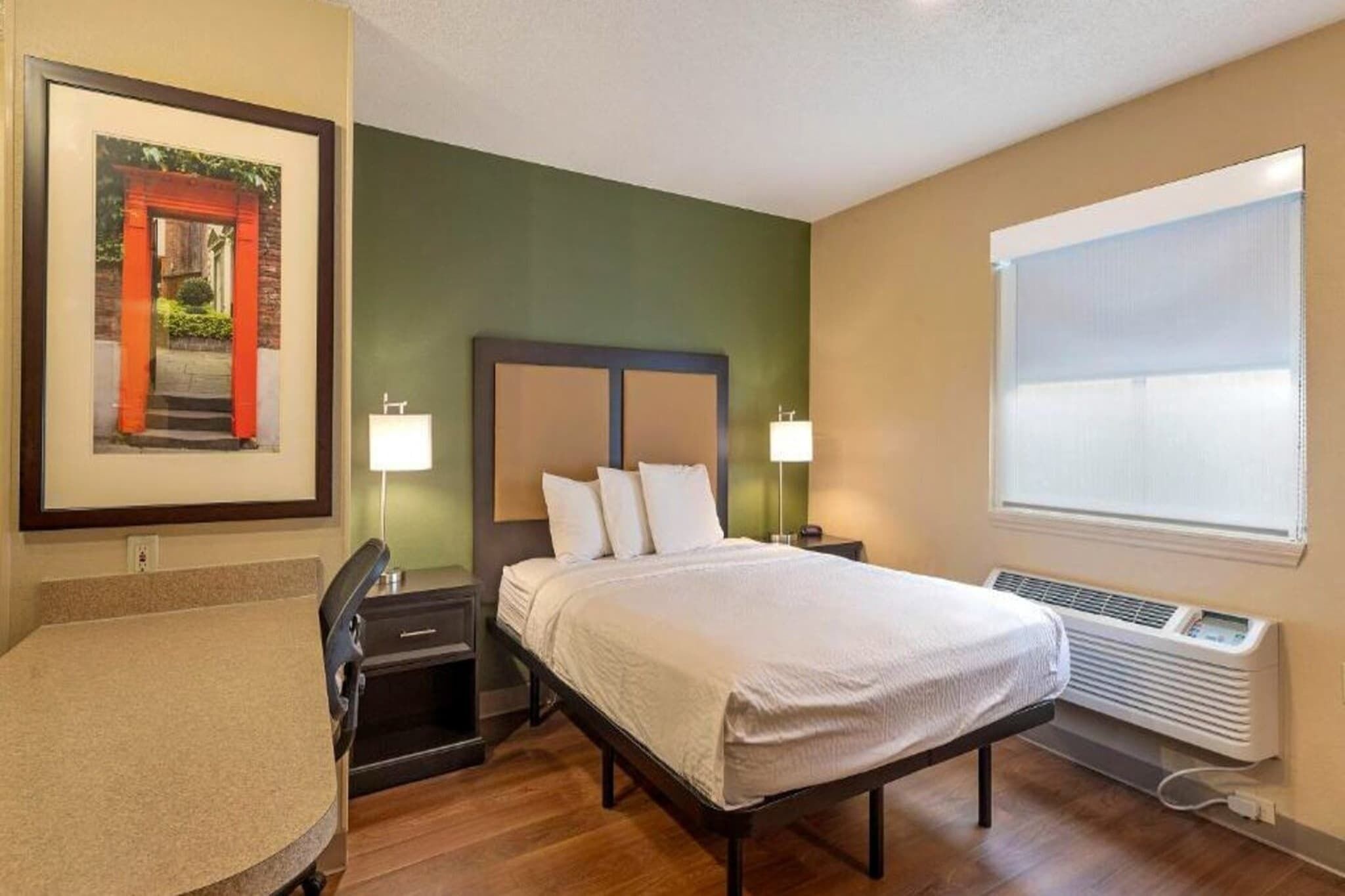 Une chambre d’hôtel comprenant un lit, des lampes de chevet, un bureau, une chaise, un tableau encadré et une fenêtre avec des stores.