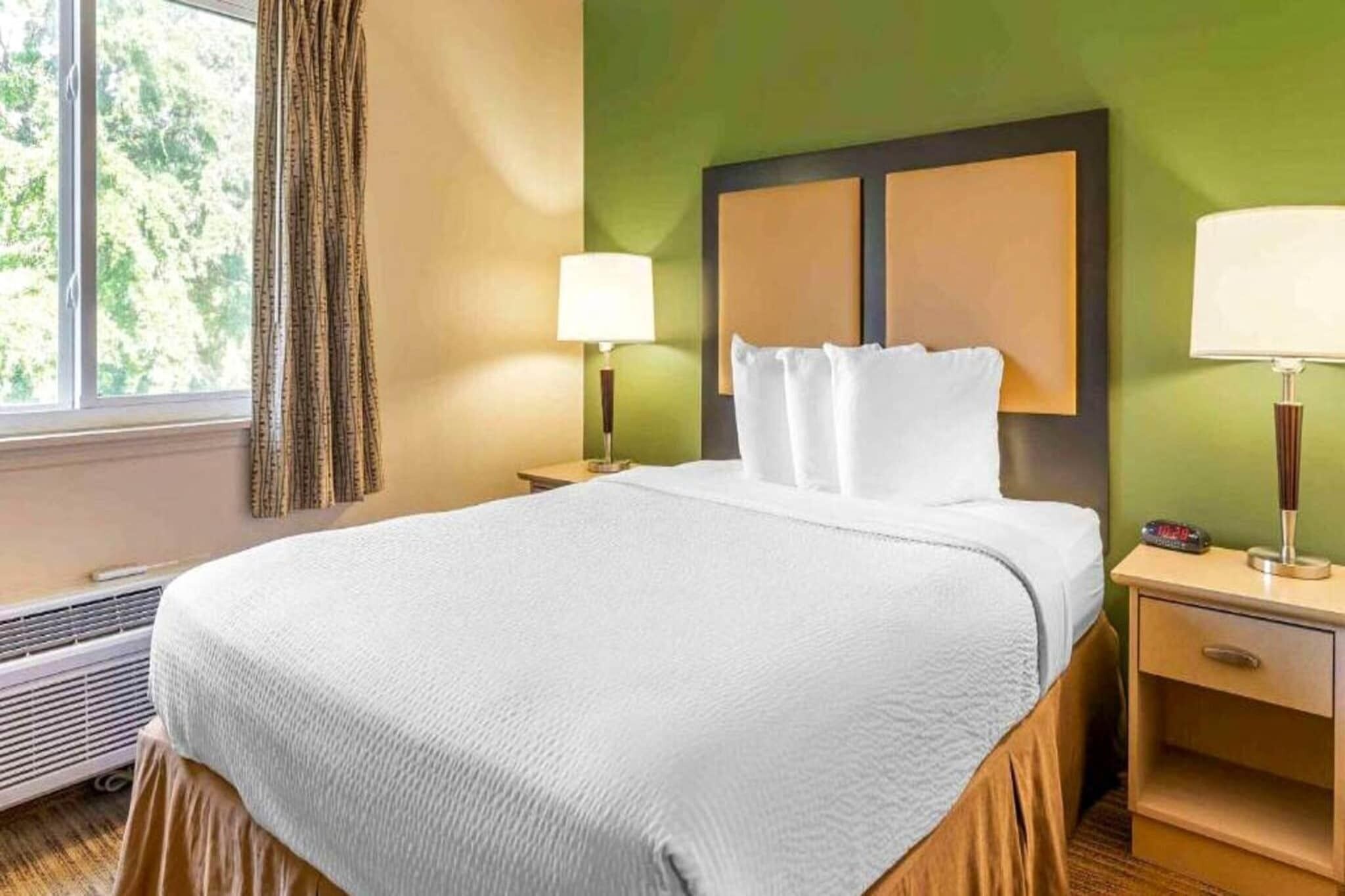 Una habitación de hotel con cama, dos lámparas de noche, una mesita de noche y una ventana con cortinas.