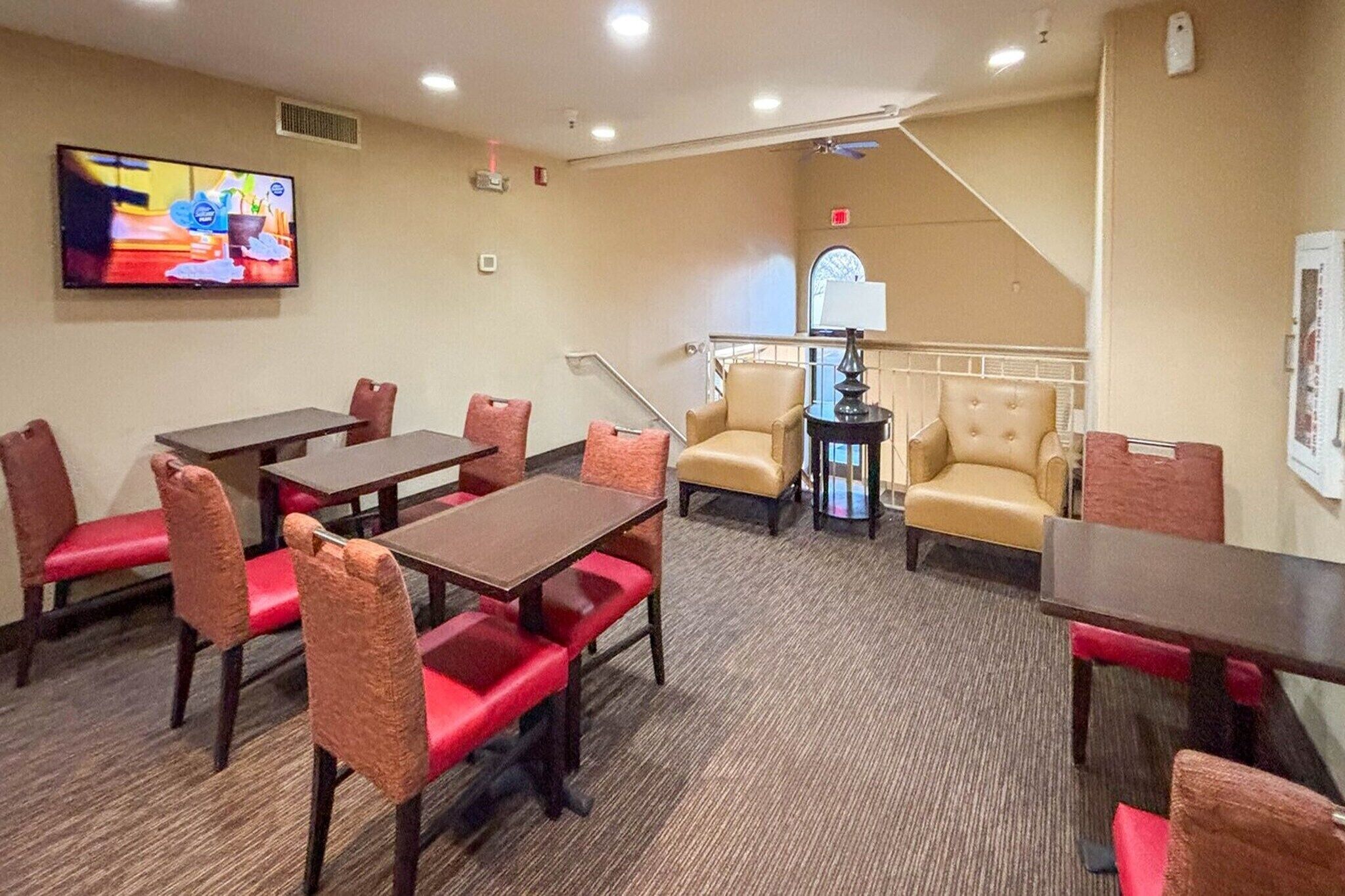 A hotel room with a dining area, a TV mounted on the wall, a staircase, and a window with an arched design.