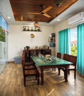 A dining area with a wooden table and chairs, a ceiling fan, and a view through a window with curtains. - The Golden View Farm & Resort (Sonkach)