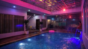 An indoor swimming pool with a glass ceiling, wooden flooring, and a seating area with chairs and plants. - The Golden View Farm & Resort (Sonkach)