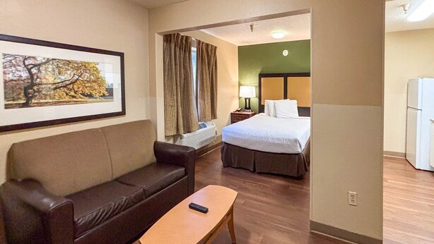 A hotel room with a bed, a sofa, a small table, a lamp, and a painting on the wall. - 1 Queen Bed | Non-Smoking, Micfridge, Sofa-Bed (Greenville SC)