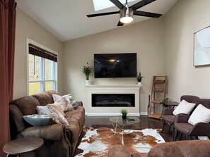 Un salon doté d’une cheminée, d’un ventilateur de plafond, d’un téléviseur à écran plat, de canapés en cuir et d’une table basse en verre.