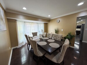 Une salle à manger avec une table en bois foncé, des chaises recouvertes d’un tissu beige et une grande fenêtre ornée de rideaux légers.