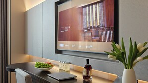A modern hotel room with a flat-screen TV mounted on the wall, a wooden dining table with a laptop, a bowl of fruit, a bottle of wine, and a vase with flowers. - Guizhou Fengrun Karst Hotel (Guiyang)