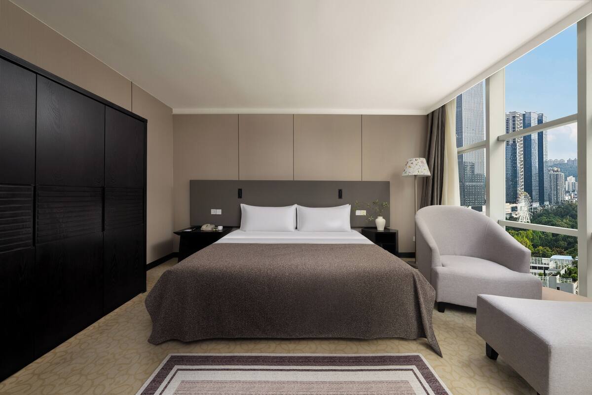 A modern hotel room with a large bed, a nightstand, a chair, and a view of the cityscape through large windows.