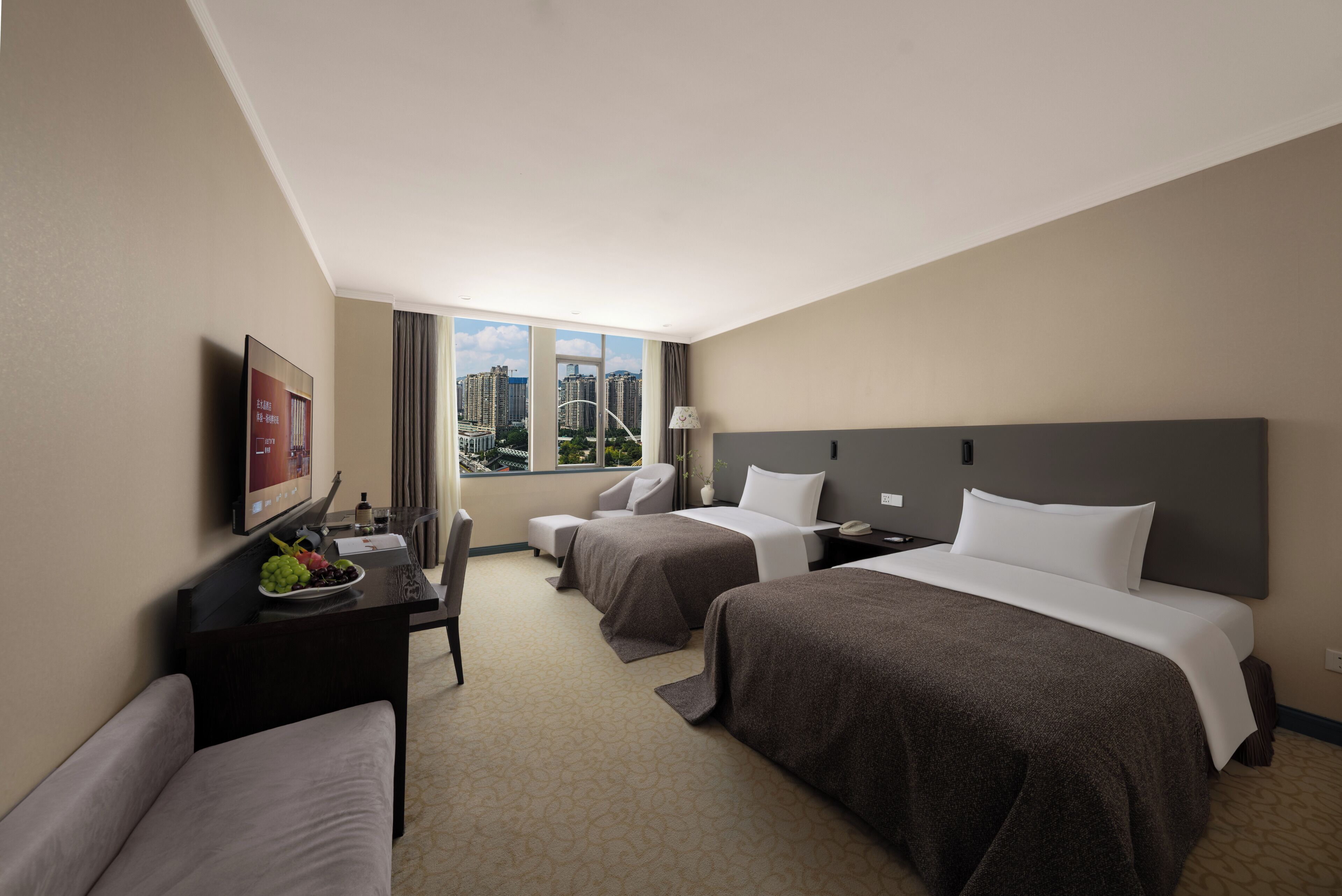 A hotel room with two beds, a desk with a computer and a plate of fruit, a chair, and a large window with a city view.