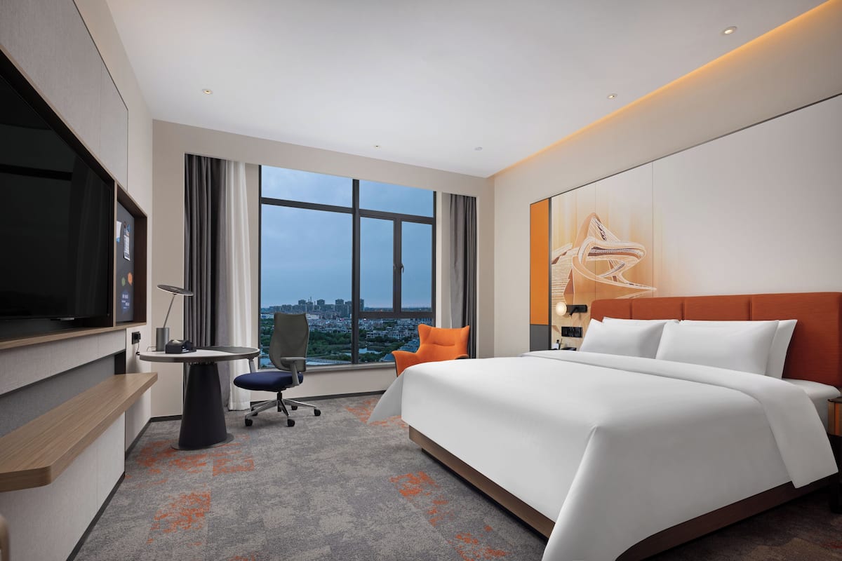 A hotel room with a large bed, a desk, a chair, a TV, and a window with a city view.