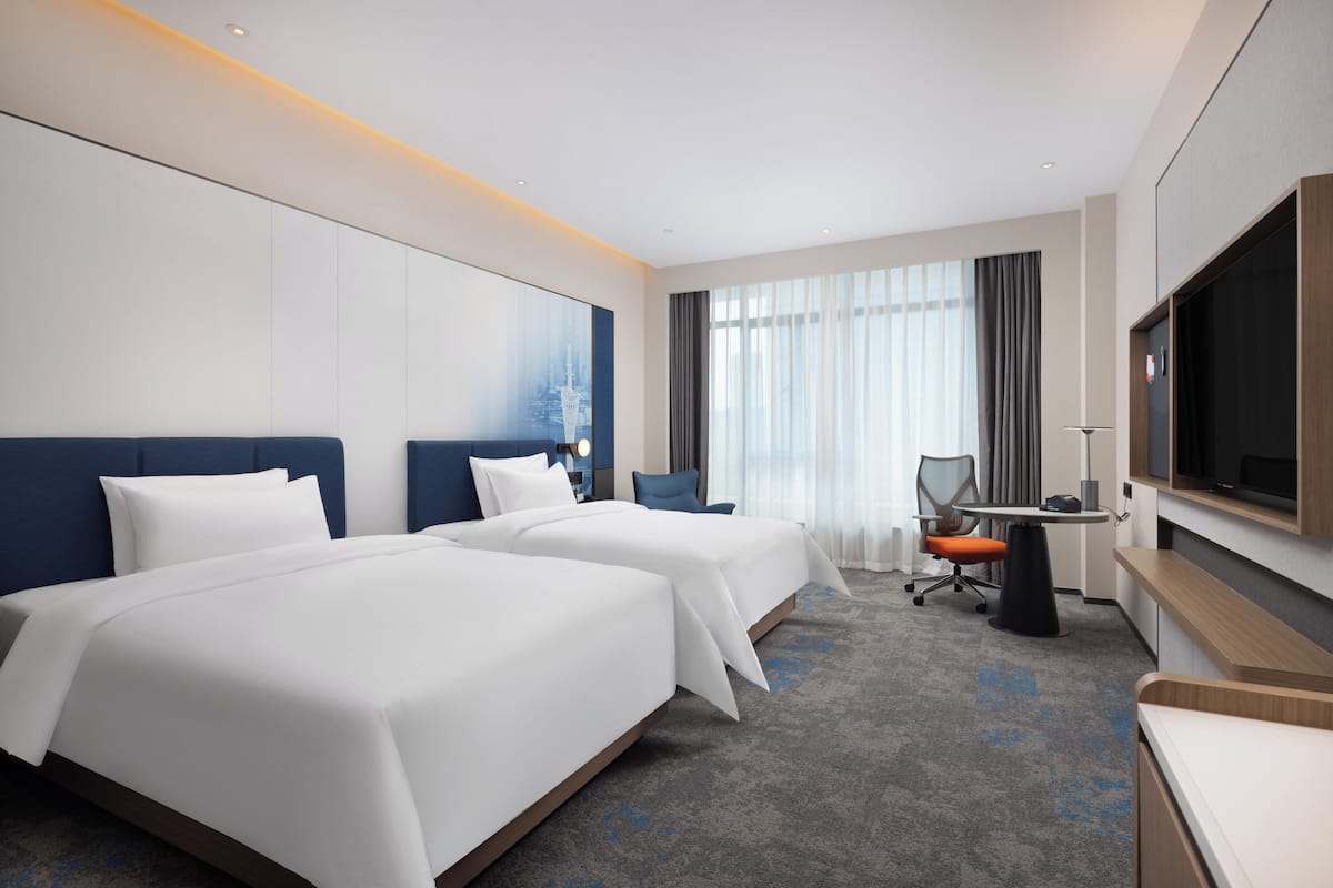 A hotel room with two beds, a desk, a chair, a TV, and a large window with curtains.