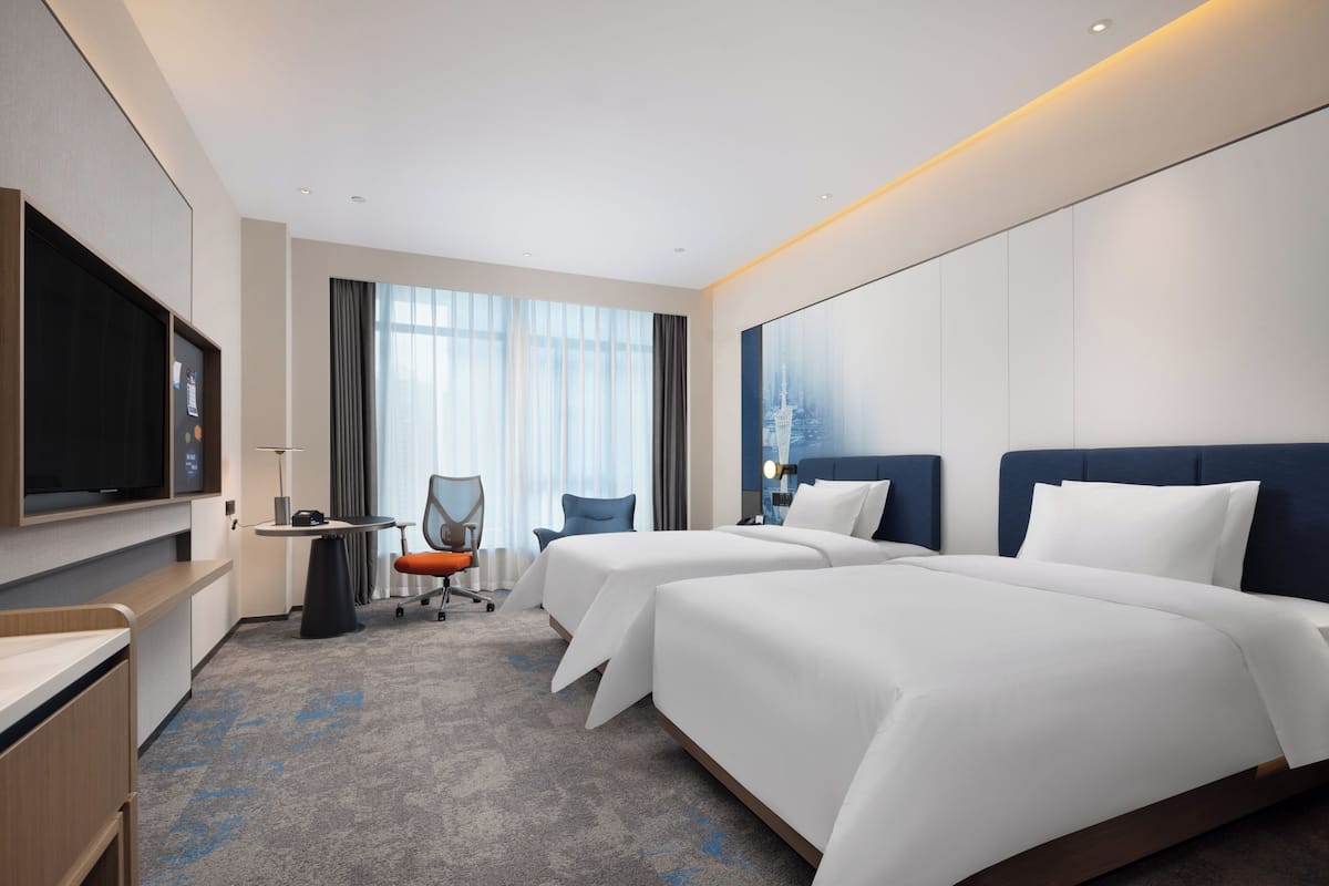 A hotel room with two beds, a desk, a chair, a TV, and large windows.