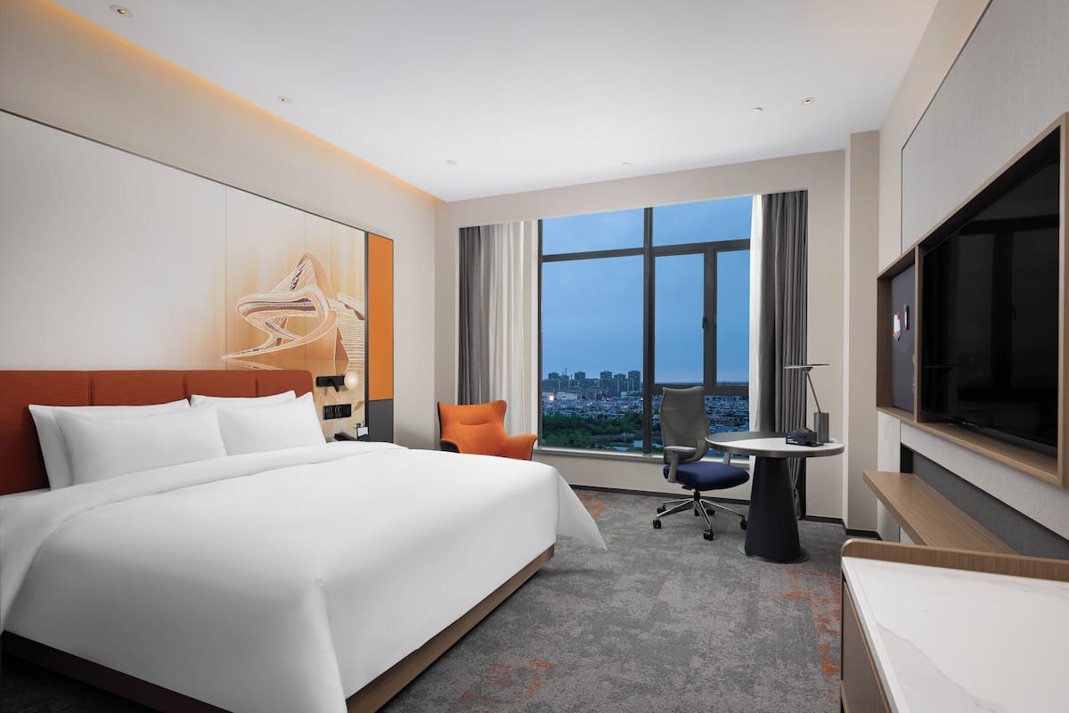 A hotel room with a large bed, a desk with a chair, a TV, and a city view.