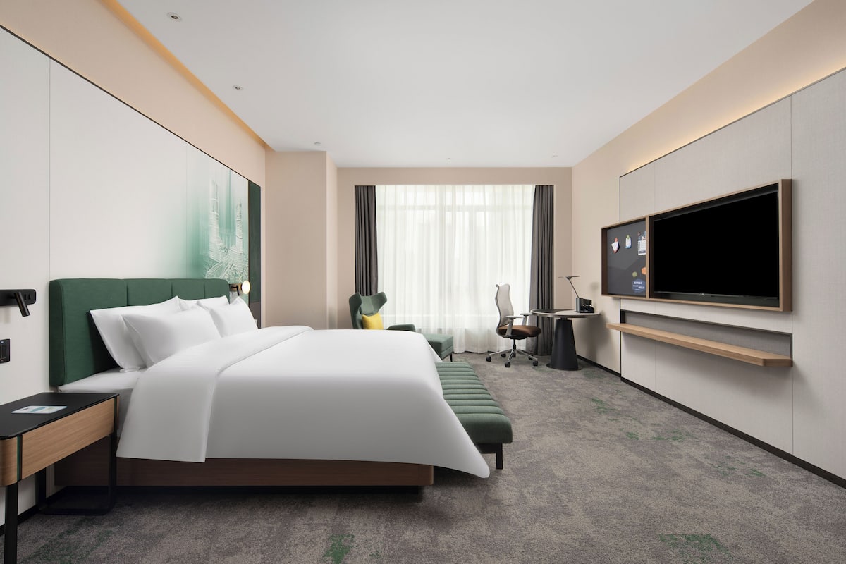 A modern hotel room with a large bed, a desk, a chair, a flat-screen TV, and a large window with curtains.