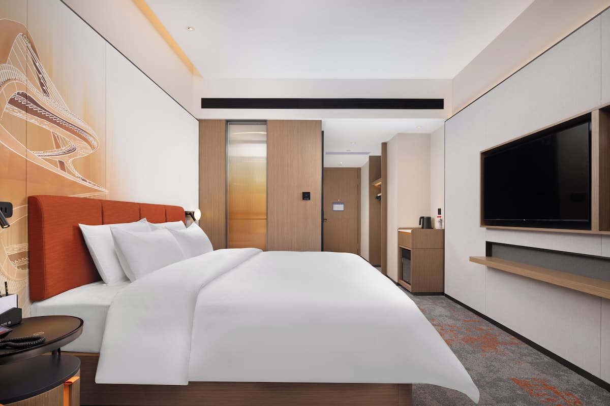 A modern hotel room with a large bed, a TV, a desk with a phone, and a bathroom in the background.