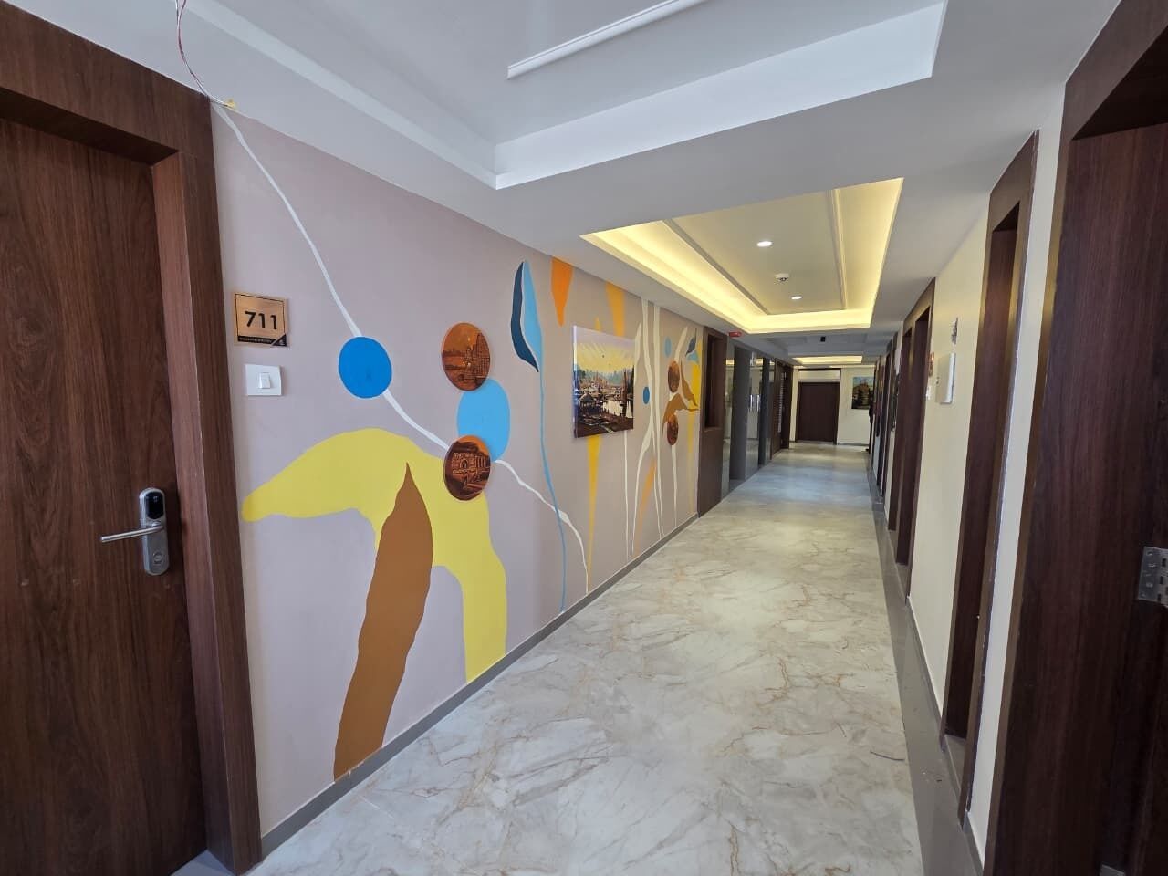 Un pasillo de hotel con piso de mármol, puertas de madera y una pared que presenta arte abstracto y un letrero con número.