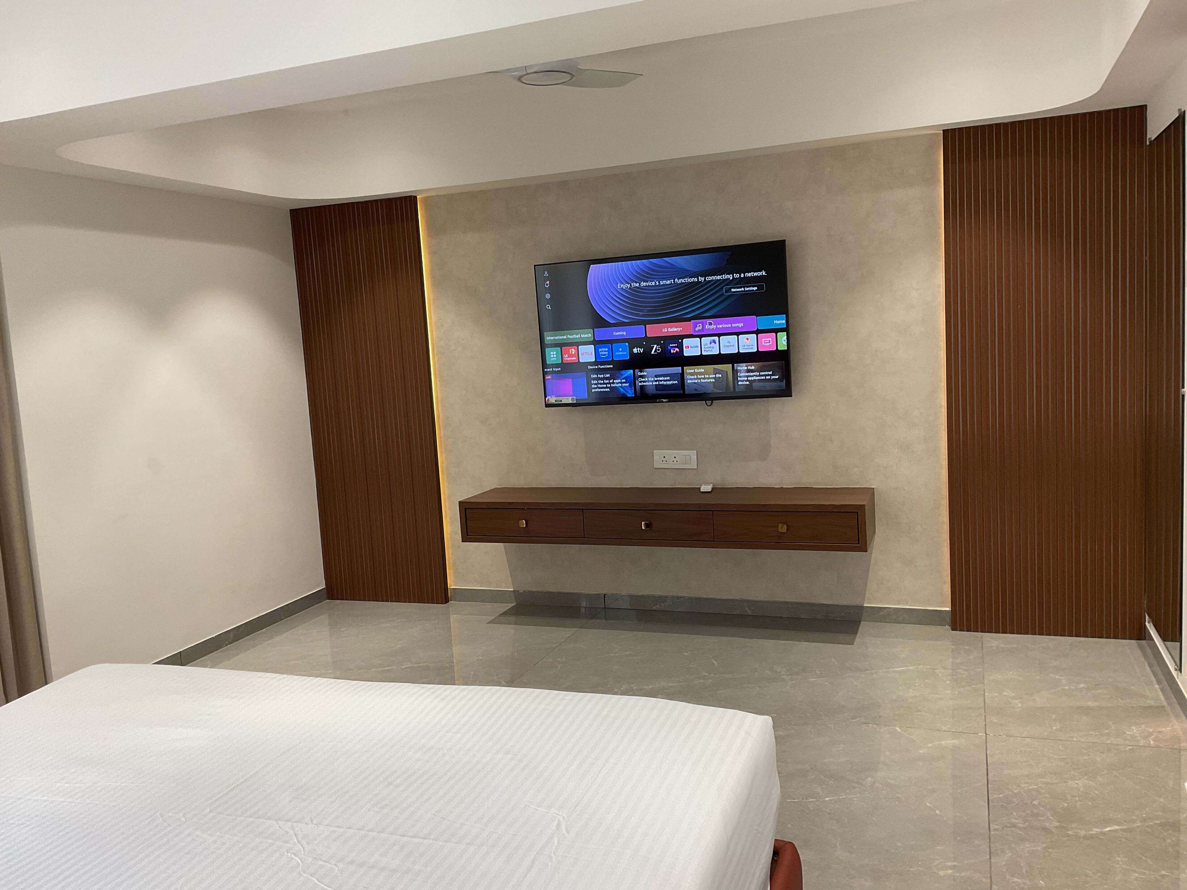 Un dormitorio moderno con un televisor de pantalla plana montado en la pared, un mueble de madera debajo y una cama con colchón blanco.
