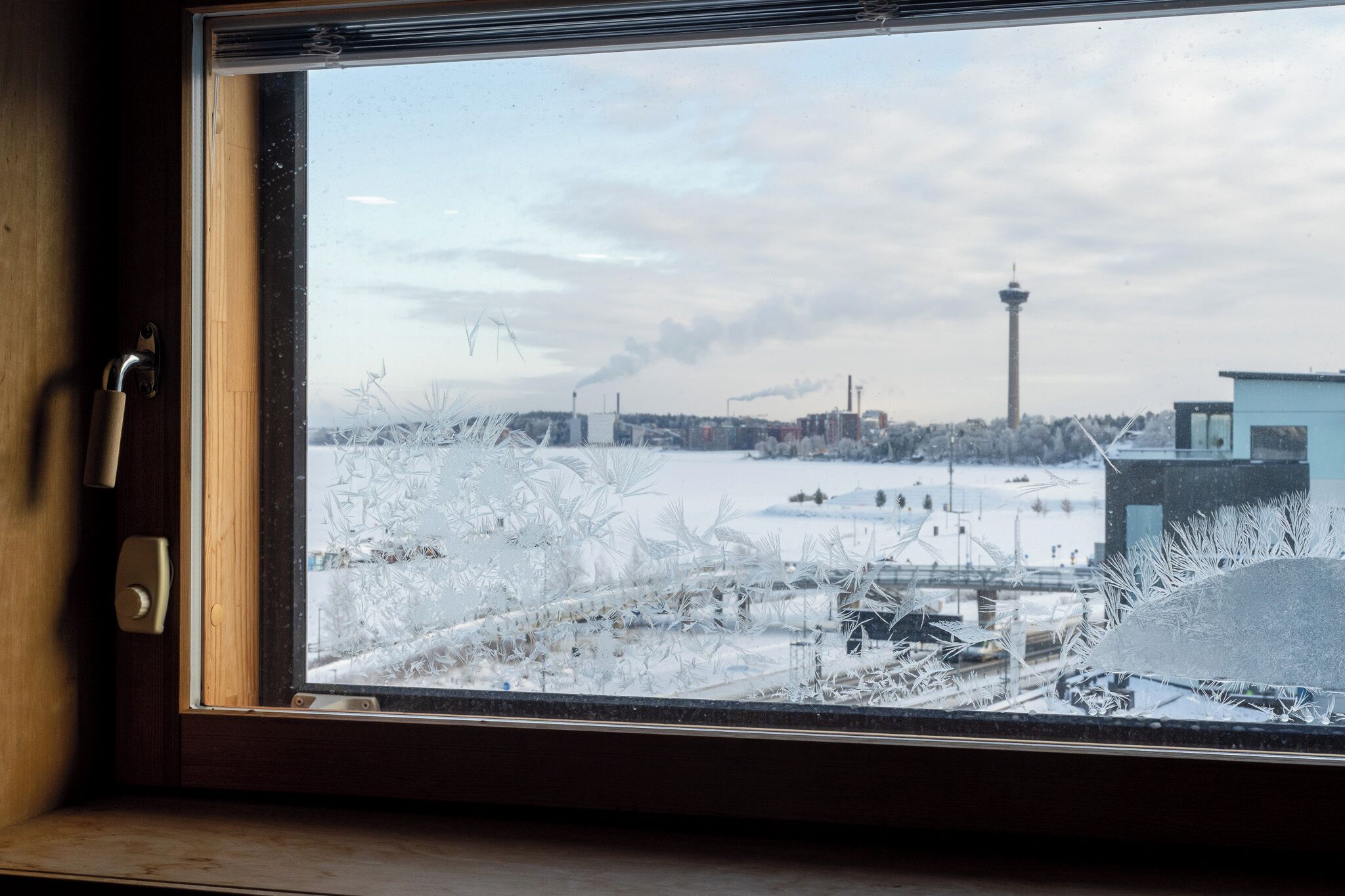 Ikkunassa näkyy huurukuvioita, ja siitä avautuu näkymä lumiseen maisemaan, ja kauempana kohoaa torni.