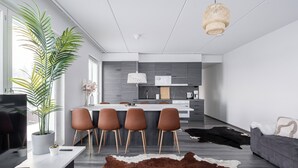 Ein modernes Wohnzimmer mit einem großen Fernseher, einer Essecke mit Stühlen, einem Sofa, einem Kuhfellteppich und einer Küche im Hintergrund.