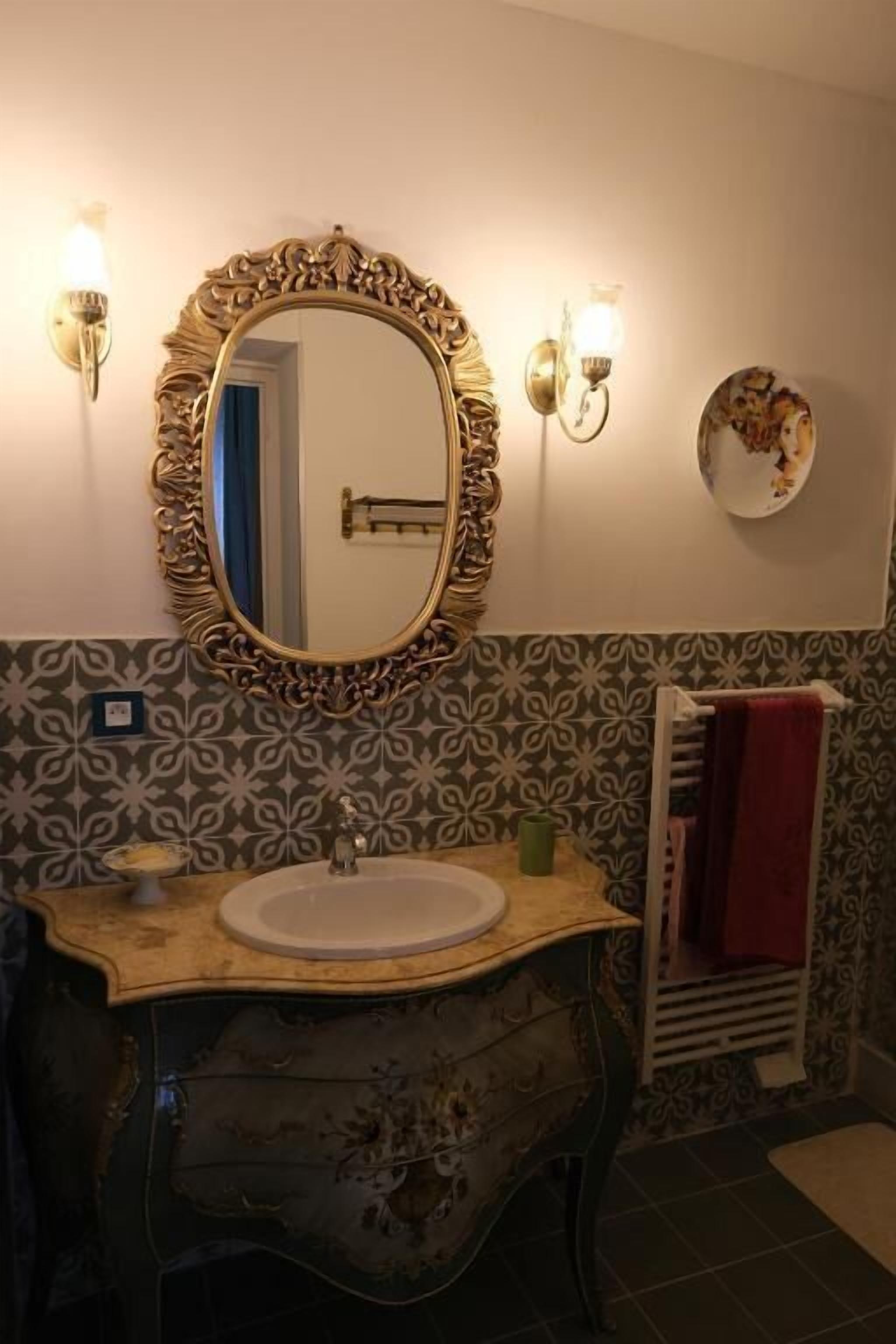 A bathroom with a large ornate mirror, a sink, a towel rack, and patterned wall tiles.