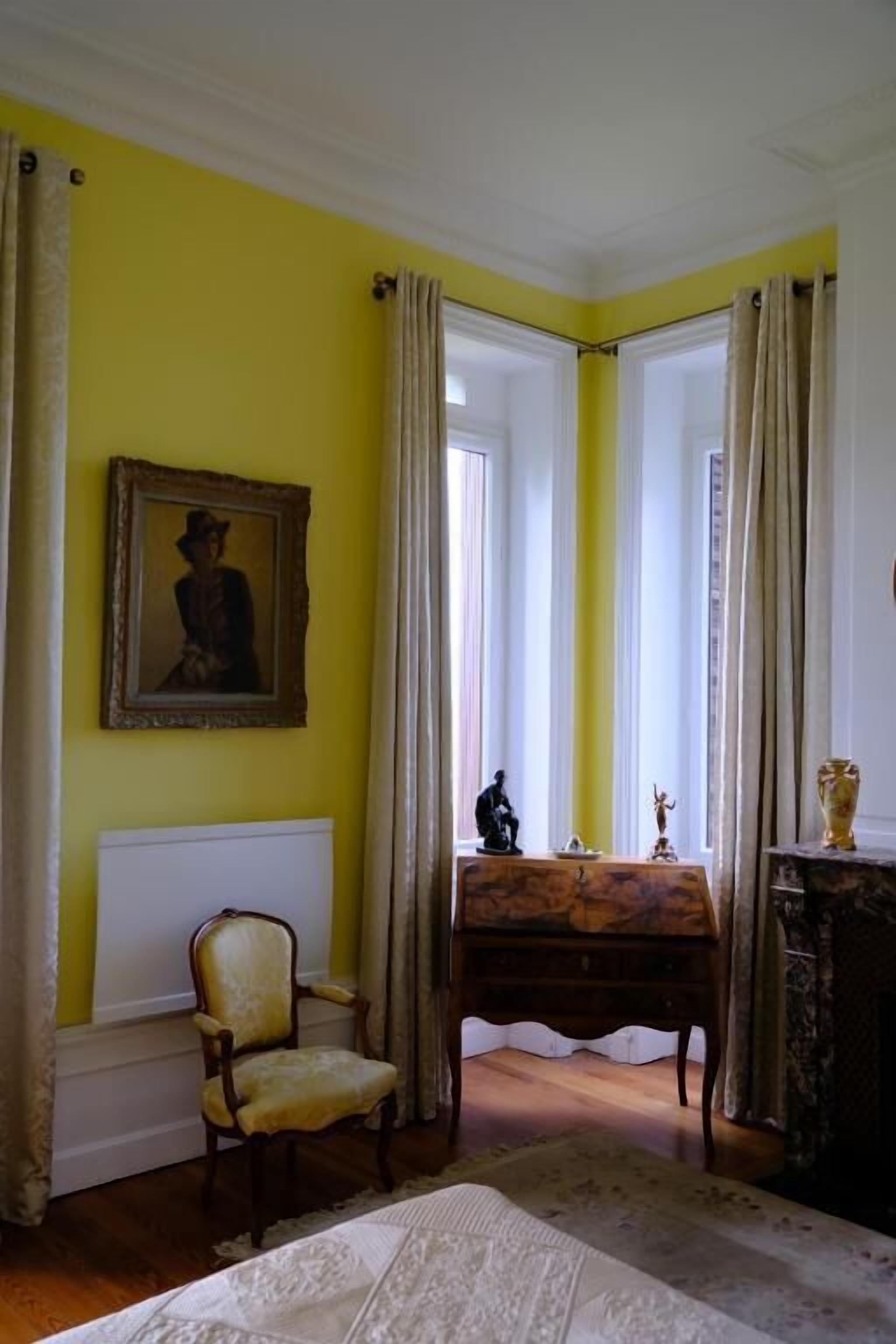 A room with yellow walls, a framed painting, a chair, a table with statues, and a window with curtains.